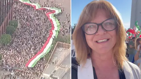 An extra long Iranian flag is carried through a crowd, with a BBC reporter smiling to camera on the right