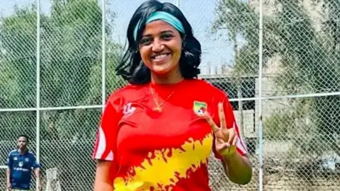 Abeba Amdu Abeba Amdu in a red and yellow football shirt holding up one her hands in a victory sign. She is standing in front of a fence at an outside sports ground. Behind her can been seen a male footballer in a navy shirt by a goal.