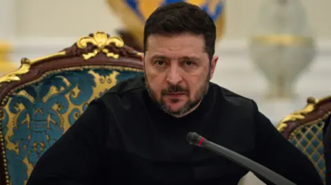 Getty Images President of Ukraine Volodymyr Zelenskyy speaks at a meeting of the National Security and Defense Council on March 3, 2026 in Kyiv, Ukraine. He's leaning forward in his chair over a microphone, with a serious look on his face, and is dressed all in black