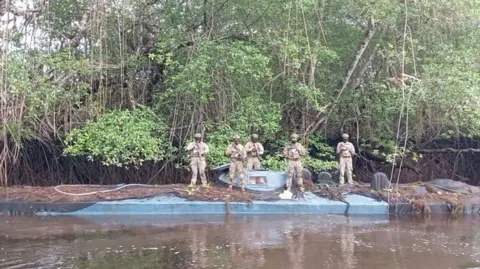 Ecuadorean Defence Ministry Five members of Ecuador's Armed Forces stand on top a semi-submersible vessel. The sub is blue in colour but parts of hit are covered by black tarpaulin and leaves and branches from the mangroves. The soldiers are armed and are in camouflage uniform and are wearing helmets. 