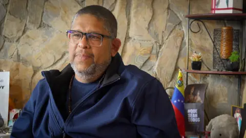 Ángel Godoy sits in a room looking serious with a stone wall behind him