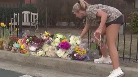 CBS News A student in shorts and a T-shirt places flowers on a pile of bouquets at a sidewalk memorial